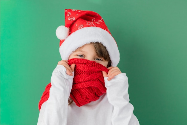 Photo bonne année et joyeux noël. jolie fille caucasienne souriante portant un bonnet de noel couvre son visage avec une écharpe rouge sur un mur vert. plaisir des vacances. copiez l'espace.