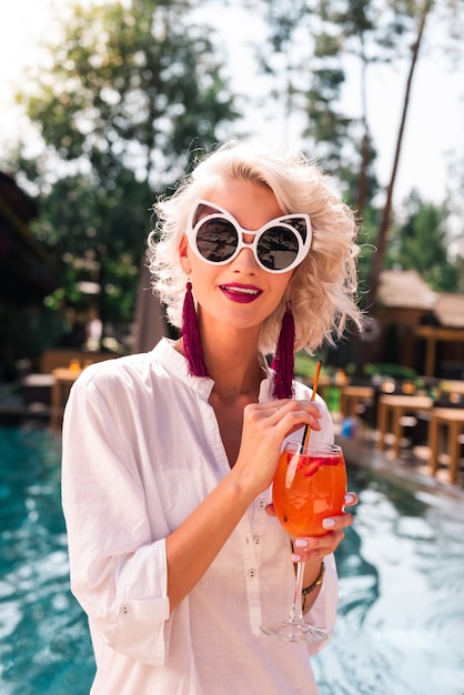 Bonne boisson. Agréable jolie femme portant des lunettes de soleil tout en buvant son cocktail près de la piscine