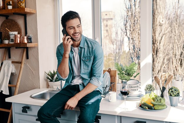 Bonne conversation. Beau jeune homme en tenue décontractée parlant au téléphone alors qu'il était assis dans la cuisine à la maison