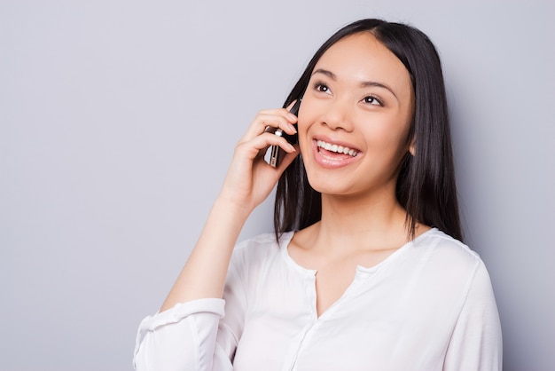 Bonne conversation. Belle jeune femme asiatique parlant au téléphone portable et souriant en se tenant debout sur fond gris