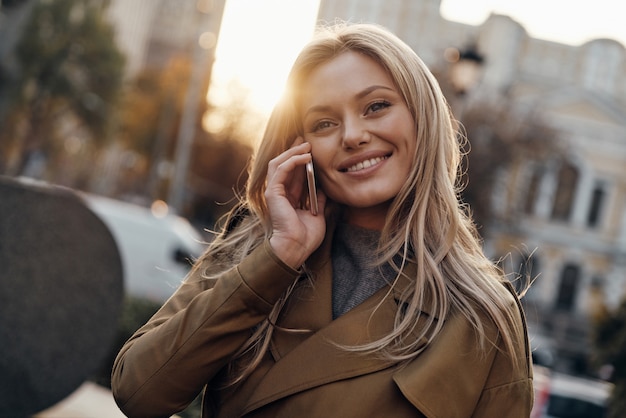 Bonne conversation. Jolie jeune femme parlant sur son téléphone intelligent et souriant tout en se tenant à l'extérieur