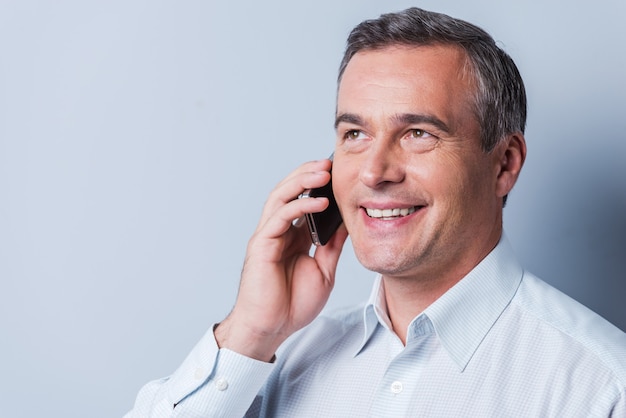 Bonne conversation. Portrait d'un homme mûr confiant en chemise parlant au téléphone portable et souriant en se tenant debout sur fond gris