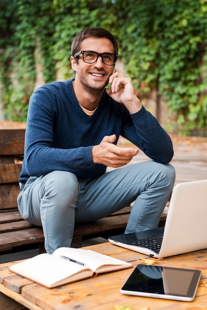 Bonne discussion d'affaires. Heureux jeune homme parlant au téléphone portable assis à la table en bois à l'extérieur