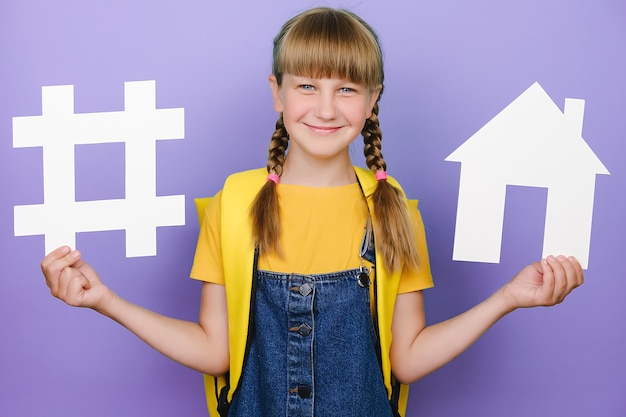 Photo bonne écolière blonde tenant un signe de hashtag blanc et un modèle de maison, étiquette pour les entreprises, porte un sac à dos jaune, posant sur fond violet. veille réseaux sociaux, mesure média, école
