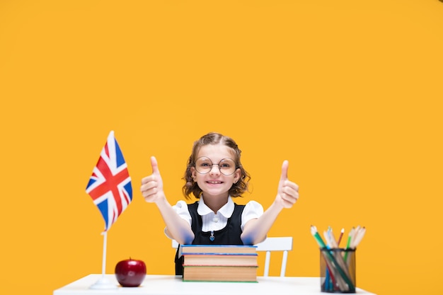 Bonne écolière caucasienne assise au bureau montrant les pouces vers le haut leçon d'anglais drapeau de la grande-bretagne