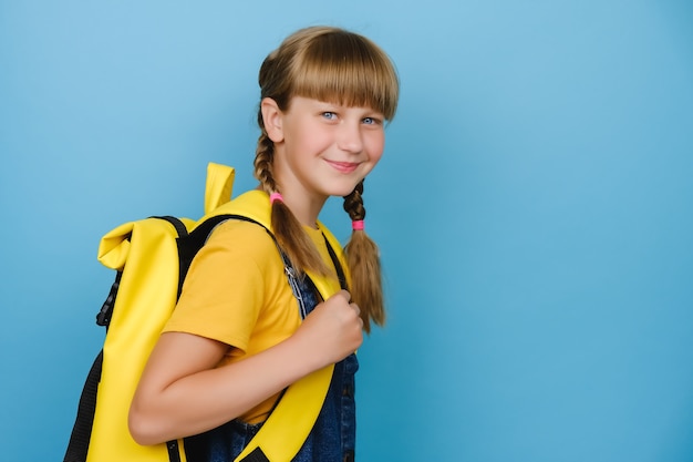 Bonne écolière souriante avec sac à dos jaune debout isolé sur fond de studio bleu avec espace de copie pour la publicité, regardant la caméra de manière positive. Portrait d'étudiant intelligent d'enfant caucasien