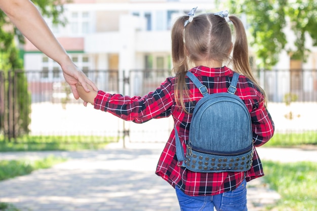 Bonne écolière tient la main de maman et va à l'école. dans un t-shirt blanc et une chemise à carreaux