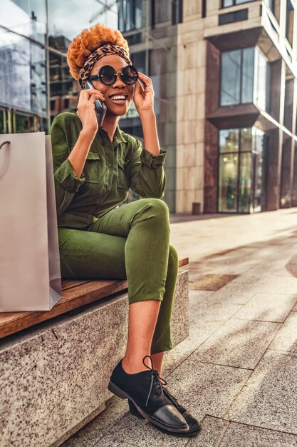 Photo bonne femme afro-américaine à la mode se détendre alors qu'il était assis sur un banc après le shopping et parler au téléphone intelligent