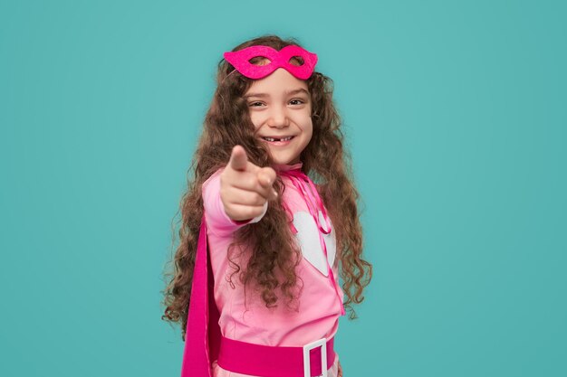 Bonne fille d'âge préscolaire aux cheveux bouclés portant un costume de super-héros rose et un masque pointant vers la caméra tout en vous choisissant sur fond turquoise