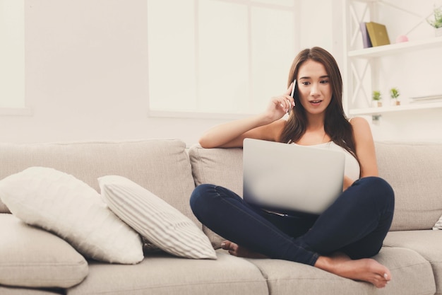 Bonne fille assise confortablement sur un canapé beige à l'aide d'un ordinateur portable, envoyant des SMS en ligne. Copier l'espace