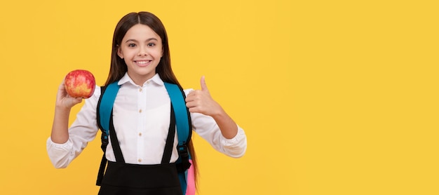 Bonne fille enfant donner le pouce en l'air tenant pomme et sac d'école fond jaune éducation Bannière d'étudiante écolière Portrait d'élève écolier avec espace de copie