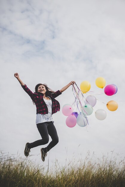 Bonne fille hipster qui saute avec des ballons en peluche colorés à l&#39;extérieur. Jeune femme s&#39;amusant dans un champ vert contre le ciel bleu. Concept de mode de vie en liberté féminine.