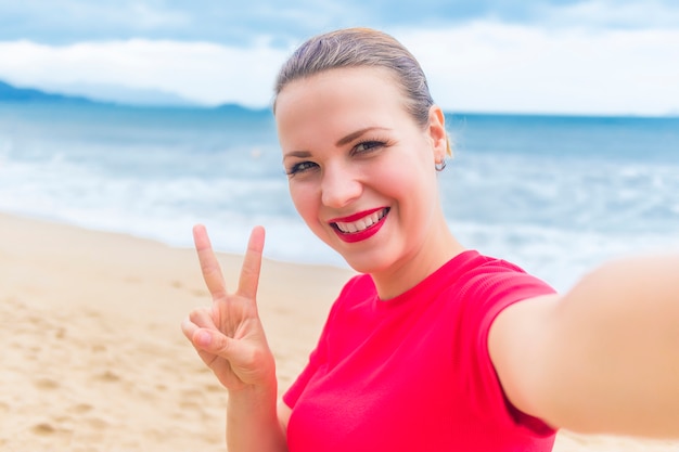 Bonne fille joyeuse positive, belle jeune femme souriante, faisant, prenant selfie, photo photo d'elle-même de vacances sur téléphone intelligent. Lady show geste de paix, signe de la victoire V.