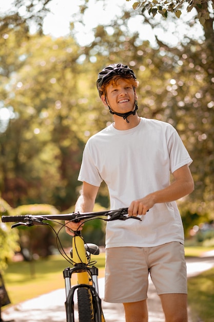 Bonne humeur. Heureux mec roux souriant en casque noir avec vélo à pied dans le parc par une chaude journée ensoleillée