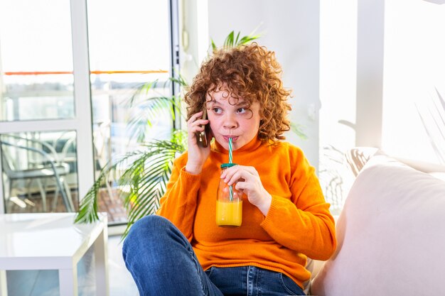 Bonne humeur jeune femme parlant au téléphone à la maison, adolescente souriante répondant à un appel par téléphone portable assis sur le canapé, ayant une agréable conversation drôle parlant par mobile et buvant du jus d'orange