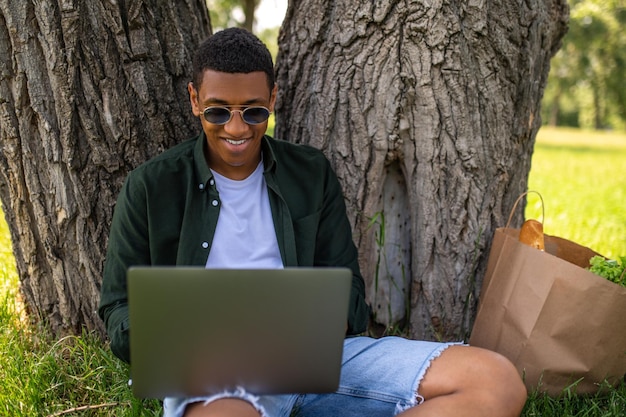 Bonne humeur. Joyeux gars à la peau foncée regardant un écran d'ordinateur portable assis près d'un arbre dans le parc le jour d'été
