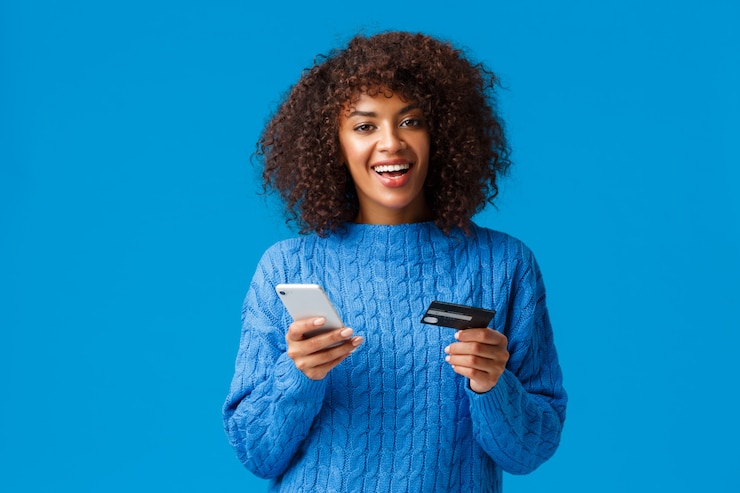 Photo bonne humeur souriante belle femme afro-américaine, acheter en ligne, faire du shopping pendant la saison des fêtes, souriant, tenant un smartphone et une carte de crédit, debout bleu