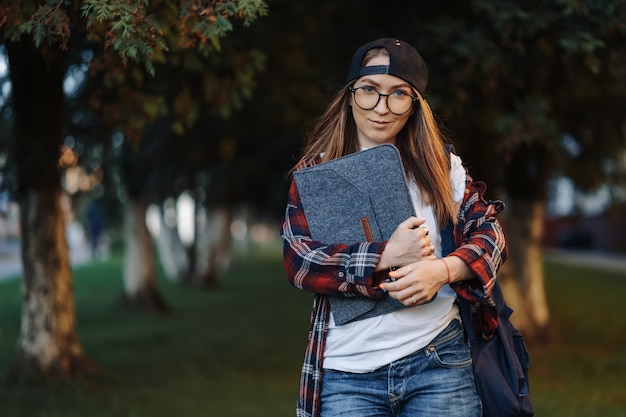 Bonne jeune étudiante vêtue de vêtements décontractés avec une tasse de café et un sac à dos derrière le dos en se promenant dans la ville. Étudiante tenant un ordinateur portable et boire du café. Heure du coucher du soleil d'été.