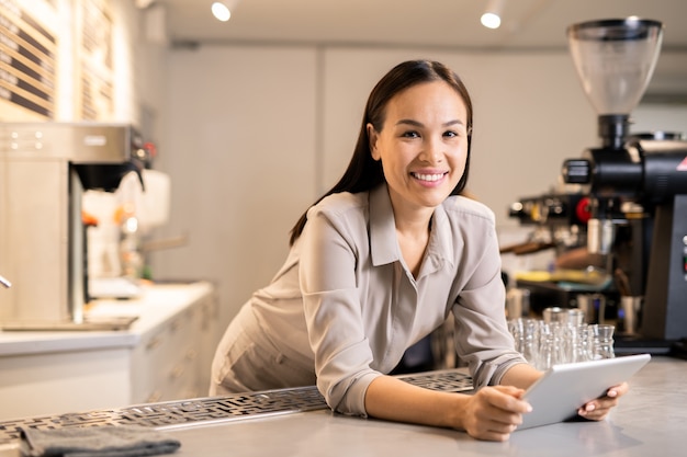 Bonne jeune femme barista avec tablette numérique en vous regardant en se tenant debout par le lieu de travail et en téléchargeant un nouveau menu pour la semaine