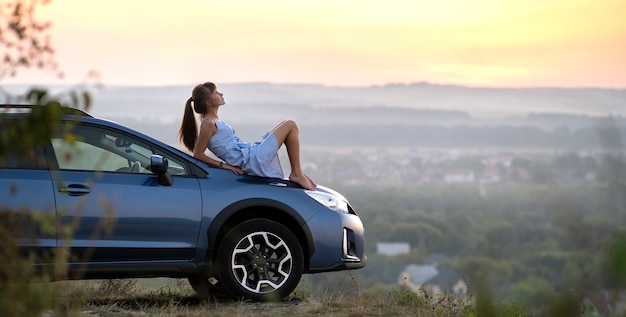 Bonne jeune femme pilote en robe bleue portant sur son capot de voiture bénéficiant d'une chaude journée d'été