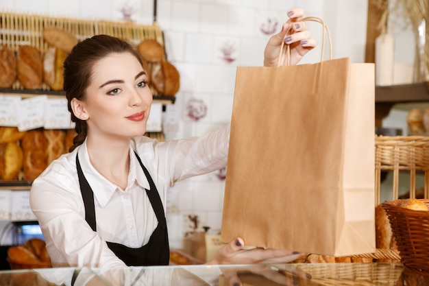 Bonne journée! Portrait horizontal d'une magnifique femme boulanger passant sac en papier avec du pain au client