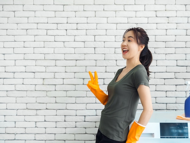 Bonne joyeuse femme asiatique souriante, femme au foyer portant des gants en caoutchouc orange montrant deux doigts, signe de la victoire près de machine à laver sur mur de briques blanches