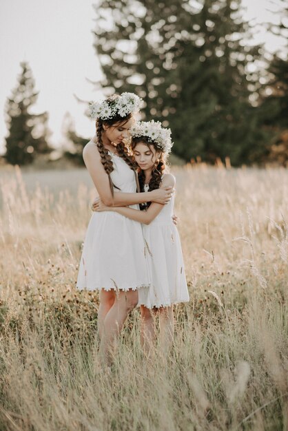 Bonne maman et sa fille en robes blanches avec des couronnes florales et des tresses style boho en été dans un champ