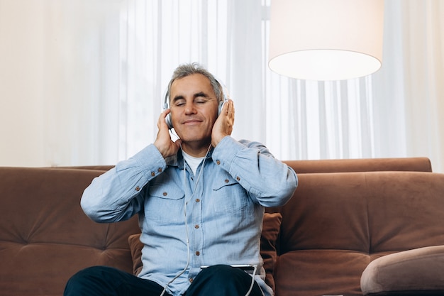 Bonne musique! Un homme d'âge moyen est assis sur un canapé marron, tient des écouteurs avec ses mains et apprécie sa musique préférée. Salon confortable sur fond. Homme en tenue décontractée utilisant un smartphone et souriant.
