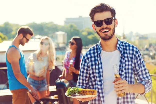 Bonne nourriture avec les meilleurs amis. Souriant jeune homme tenant une bouteille avec de la bière et une assiette avec de la nourriture tandis que trois personnes font un barbecue en arrière-plan