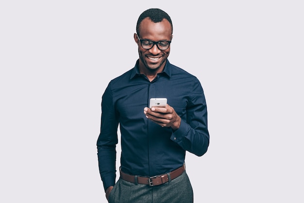 Bonne nouvelle d'un ami. Beau jeune homme africain tenant un téléphone intelligent et le regardant avec le sourire en se tenant debout