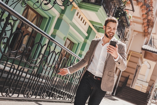 Photo bonne nouvelle homme brun souriant debout à l'extérieur et utilisant son smartphone, il tient son téléphone