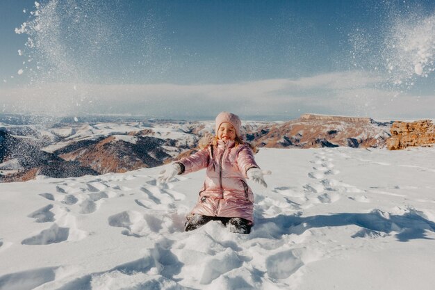 Bonne petite fille assise dans la neige et jetant de la neige avec joie. notion d'hiver