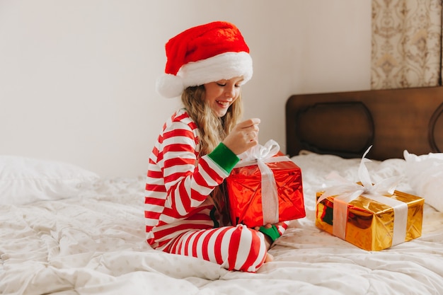 Bonne petite fille au chapeau de père Noël est assise sur le lit avec un cadeau de Noël. Enfant de Noël, enfant.