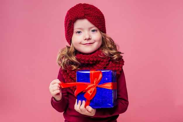 Bonne petite fille blonde souriante aux cheveux bouclés en bonnet rouge tricoté avec boîte-cadeau de Noël avec archet sur fond rose studio. Nouvel An présent dans les mains d'une enfant de sexe féminin faisant un voeu copyspace