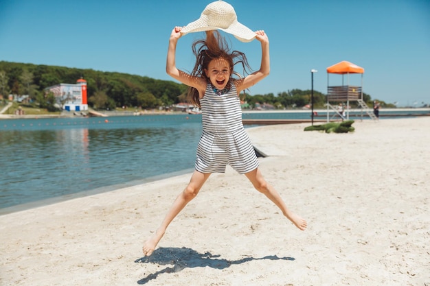 Bonne petite fille dans un chapeau de paille sautant sur la plage en vacances d'été