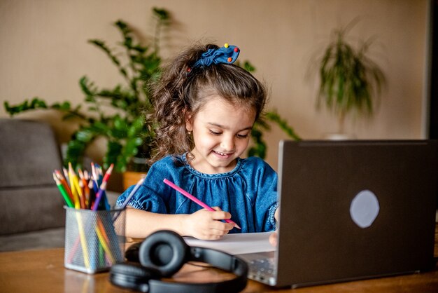 Bonne petite fille dans les écouteurs en souriant et en prenant des notes dans le bloc-notes tout en communiquant avec l'enseignant par vidéo