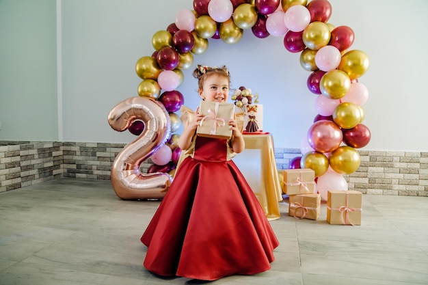 Bonne petite fille de deux ans tenant son cadeau d'anniversaire devant son gâteau d'anniversaire décoré de ballons multicolores. Belle séance photo d'anniversaire de petite fille