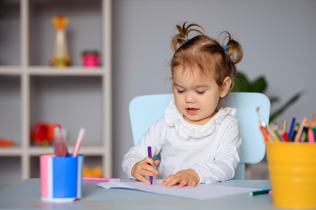 Bonne petite fille est assise à la table et dessine sur papier avec des crayons de couleur