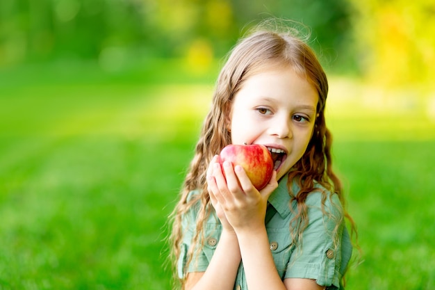 Bonne petite fille en été sur la pelouse mord une pomme rouge avec des dents saines sur l'herbe et sourit de l'espace pour le texte