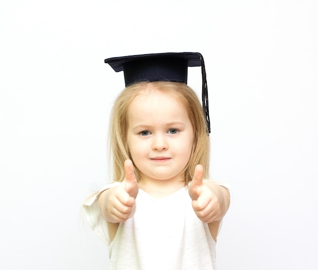 Bonne petite fille étudiante au chapeau étudiant montrant les pouces vers le haut en fond blanc isolé