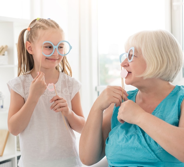 Bonne petite fille avec grand-mère mettant des lunettes et des lèvres sur des bâtons dans une pièce lumineuse