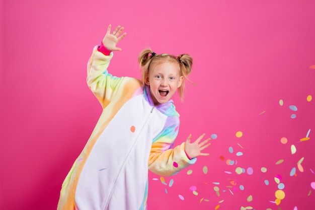 Bonne petite fille en licorne kigurumi sur un mur rose se réjouit de confettis multicolores