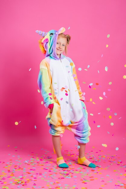 Bonne petite fille en licorne kigurumi sur un mur rose se réjouit de confettis multicolores