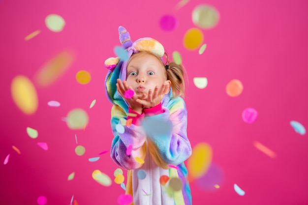 Bonne petite fille en licorne kigurumi sur un mur rose se réjouit de confettis multicolores