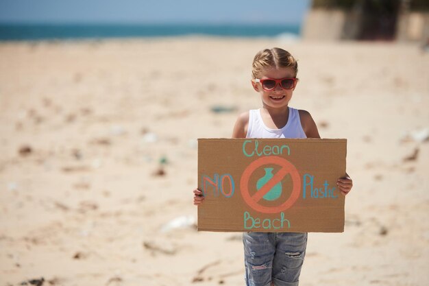 Bonne petite fille à lunettes de soleil tenant une pancarte en carton essayant d'organiser le nettoyage de la plage