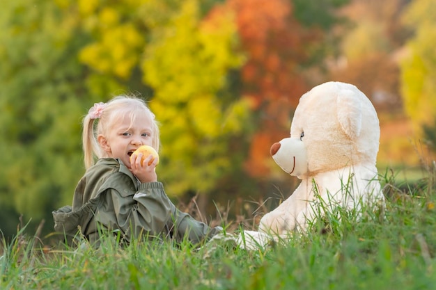 Bonne petite fille mange une pomme et joue avec un grand ours en peluche assis sur l'herbe Enfant le jour de l'automne dans la nature