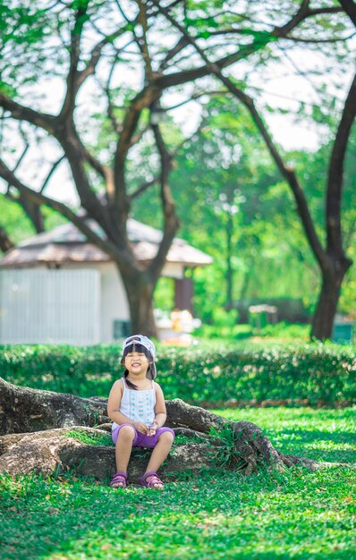 Bonne petite fille porter un chapeau assis sur la racine de l&#39;arbre dans le parc