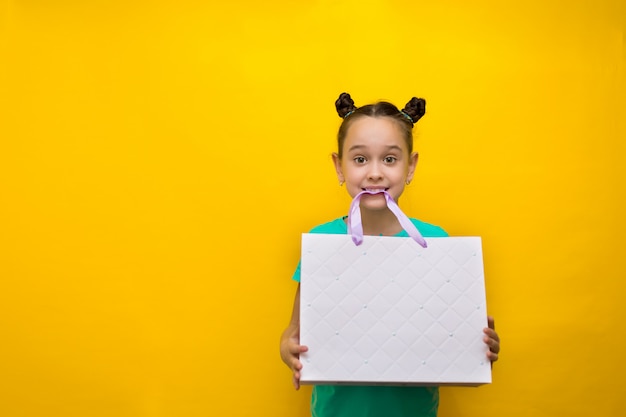 Bonne petite fille avec des queues drôles debout isolé sur un mur jaune tenant le paquet dans ses dents. Regarder la caméra