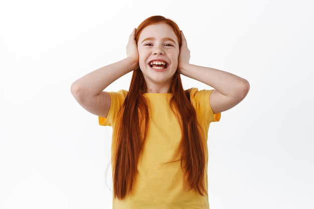 Bonne petite fille rousse avec des taches de rousseur riant, souriant et couvrant les oreilles avec les mains, ne voulant pas écouter, jouant, debout sur fond blanc