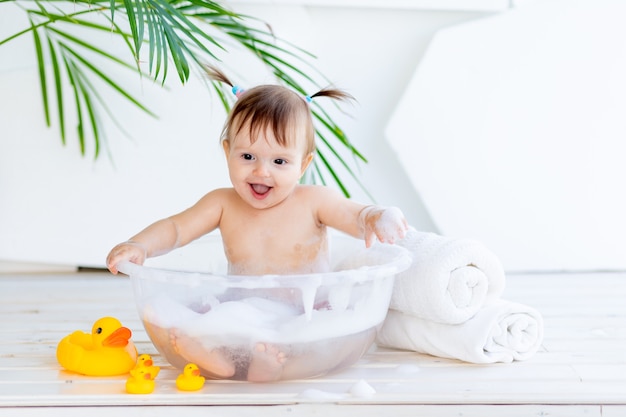 Bonne petite fille se lave et joue dans un bassin avec de la mousse et de l'eau dans une pièce lumineuse à la maison